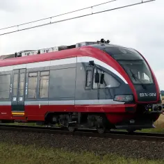 Czerwono-szary pociąg elektryczny ED74-001d PKP Intercity na torach, z polami w tle.